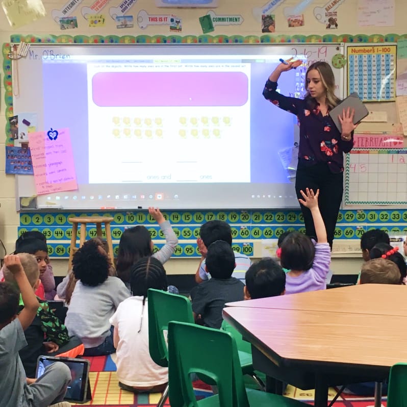 Photo of a teacher standing in front of a classroom of students raising their hands